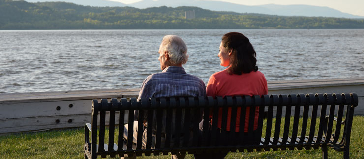 Elderly woman and daughter sit on bench quietly looking at the river on a sunny day.