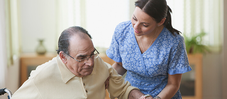 Senior man in wheelchair gets help standing up from a female care aide.