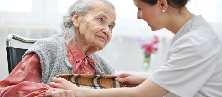 Female home care worker places a blanket on an elderly woman sitting in a wheelchair at home.