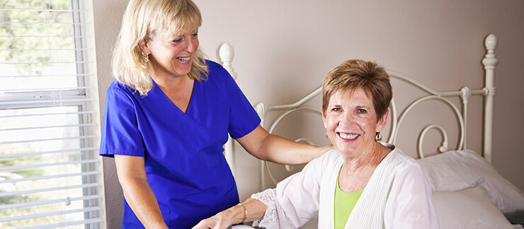 Senior woman sitting in bed smiles as female caregiver comes for a visit.