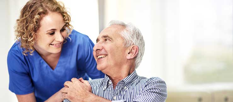 Female care provider smiles while holding the hand of a male senior care recipient.