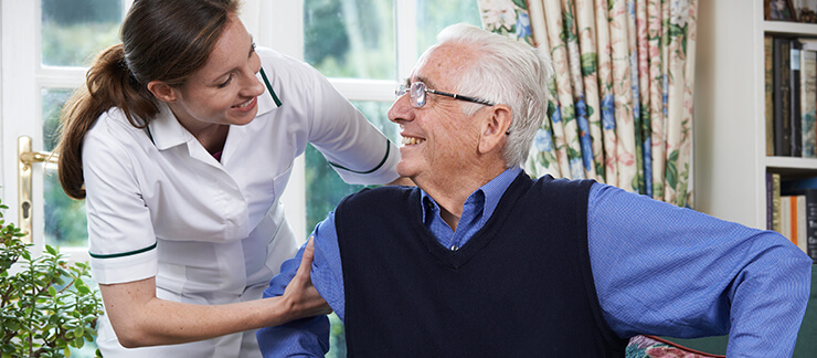 Female care worker helps senior man with paranoia get up from sitting on couch.