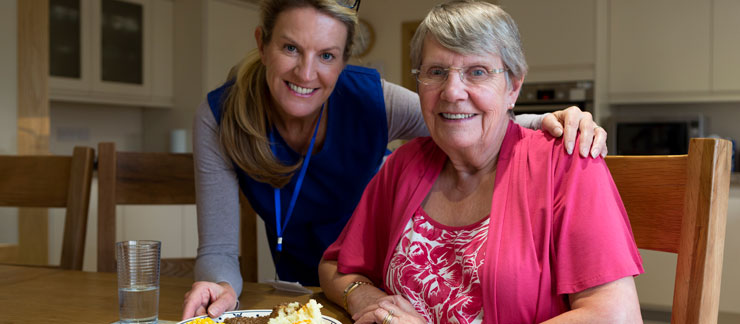 In-home care provider leaning over at a kitchen table with her arm around an elderly woman in pink