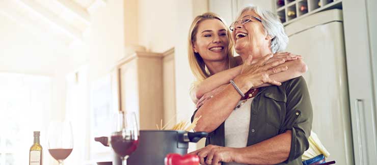 Senior woman and her adult daughter laugh while cooking pasta in the kitchen.