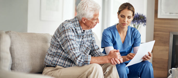 Senior man sits on a couch listening to female caregiver read to him from a paper form.