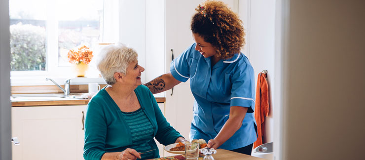 Professional caregiver serves lunch to senior woman sitting in kitchen.