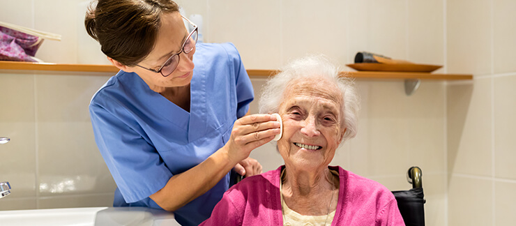 Elderly woman gets face cleaned in bathroom by home care aide.