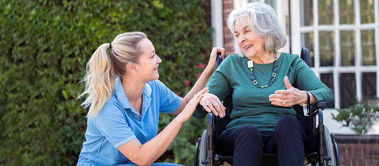 Female personal care worker kneels down next to senior woman in wheelchair outside of her home.