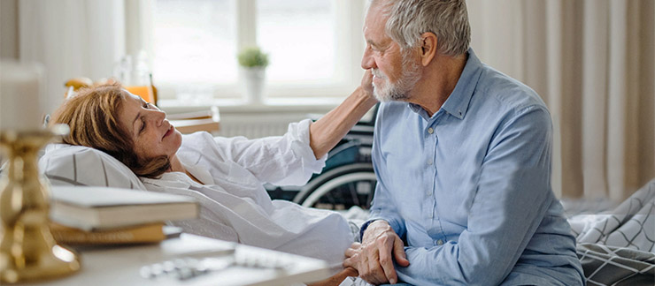 Husband holds hand of ill wife as she lays in bed at home.