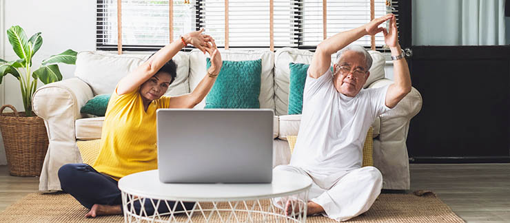 Elderly couple sitting on the floor in front of computer while doing stretching exercises at home.